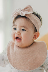 Baby wearing Avauma Mellow Ribbon Band in Baby Blush with matching pink bib, smiling